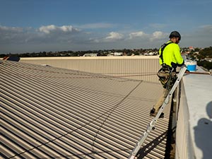 Standing-Seam-Metal-Roof-Elizabethtown-KY-Kentucky-1 | Commercial Roofing Pros Worker in a high-visibility vest and helmet stands on a corrugated metal roof, secured with a harness near the edge on a sunny day.