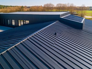 standing-seam-metal-roof-Elizabethtown-KY-Kentucky-2 | Commercial Roofing Pros Blue corrugated metal roof viewed from above, with a small vent and surrounding low building; fields and trees in the background.