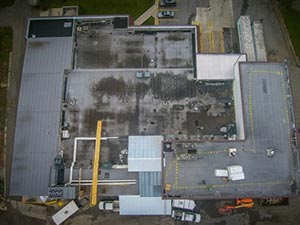 Aerial view of a large industrial building roof with HVAC units, piping, and a yellow crane running across the top.